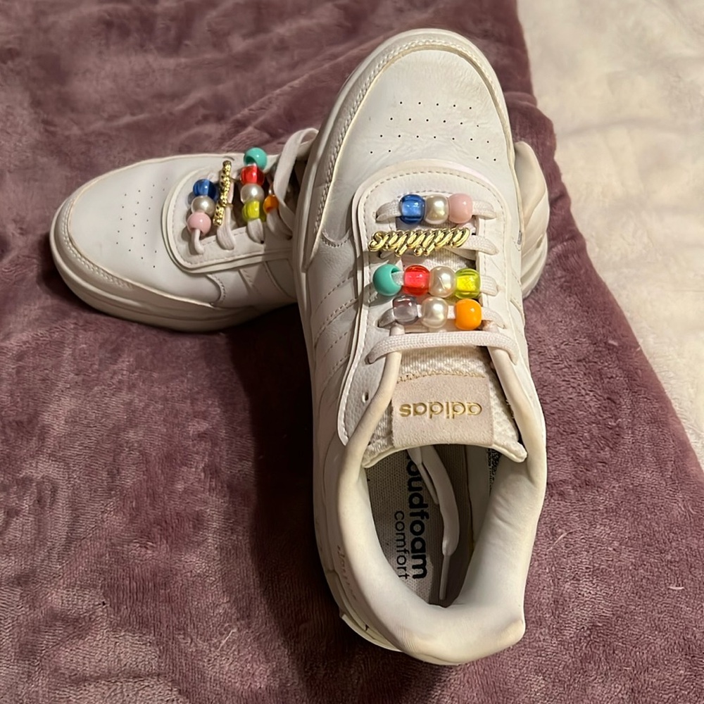 White Adidas shoes with colored beads and emblem on laces.